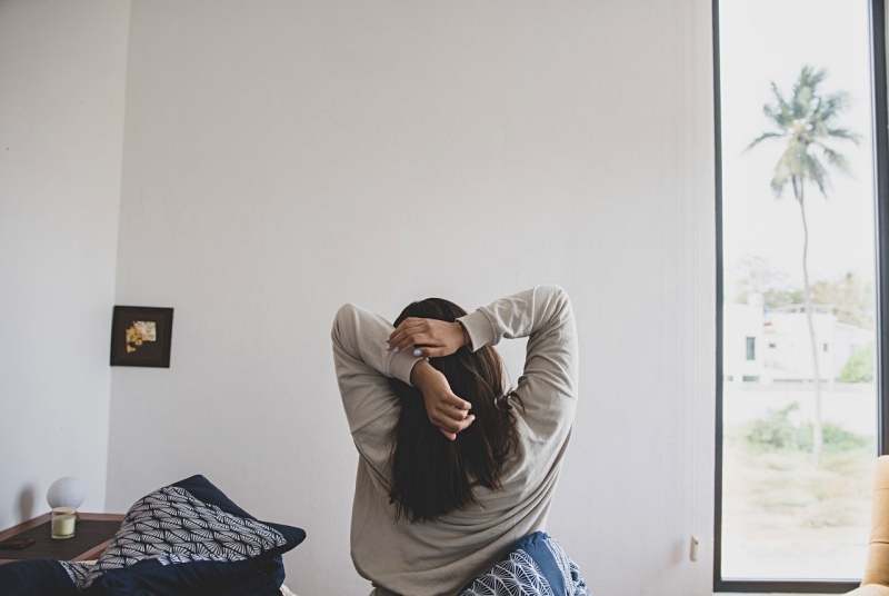 a back view of a woman in gray sweater stretching her arms in the morning | Tips On How To Wake Up Early | Featured