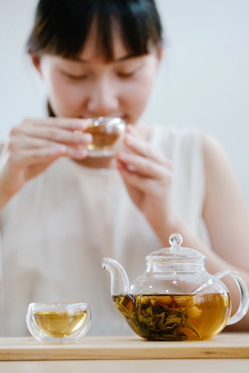 glass teapot on a table and woman sipping tea from a small glass | natural herbs